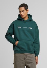 Sudadera verde oscura con un bolsillo frontal, con logotipos de texto en blanco. Hecha de un material suave, tiene un corte relajado y capucha ajustable con cordón.