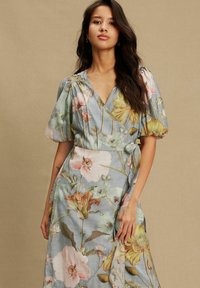 Floral Wickelkleid aus hellblauem Stoff. Mit Puffärmeln, tiefem V-Ausschnitt und einem Taillengürtel. Das Muster zeigt große, bunte Blumen.