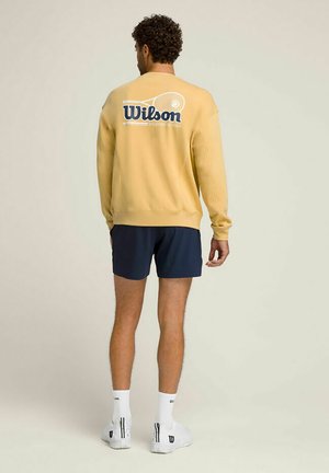 Mann trägt gelben Wilson-Sweatshirt mit Tennisracket-Grafik, marineblaue Shorts, weiße Socken und weiße Turnschuhe, steht mit dem Rücken zur Kamera vor einfarbigem Hintergrund.