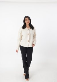 Cardigan color crema, suave, con cuello, que presenta cuatro botones beige; combinado con pantalones negros y sandalias con tacones transparentes.