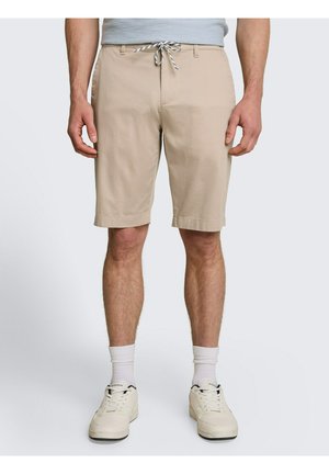 Mann trägt beige knielange Shorts mit Kordelzug in der Taille, weiße Socken und weiße Turnschuhe, steht vor einem schlichten hellen Hintergrund.