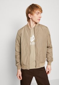 Beige Bomberjacke mit Reißverschluss vorne, gerippten Bündchen und Saum. Getragen über einem hellen T-Shirt mit grafischem Design.