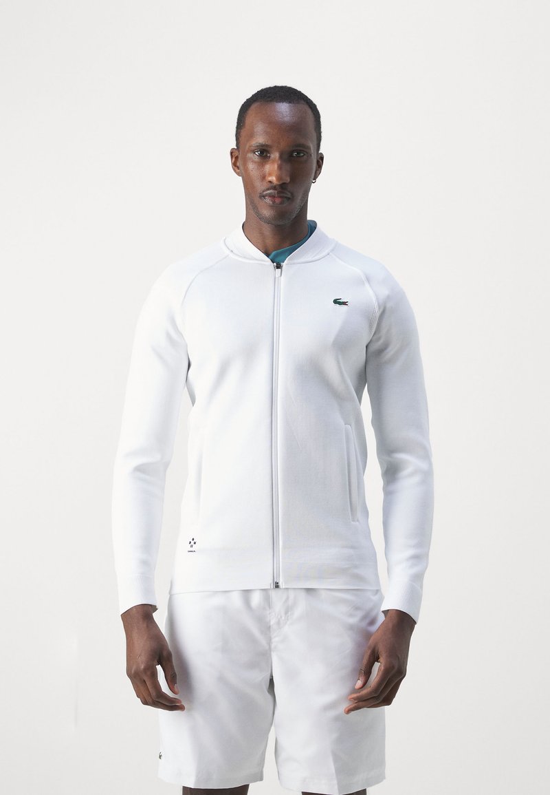 Sweat-shirt zippé blanc avec des manches longues et un col côtelé, arborant un petit logo vert sur la poitrine et deux poches avant.