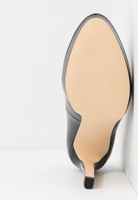 Semelle d'une chaussure à talon noir inclinée contre une surface blanche, montrant une semelle beige avec une texture antidérapante et le nom de la marque "GUESS" embossé.