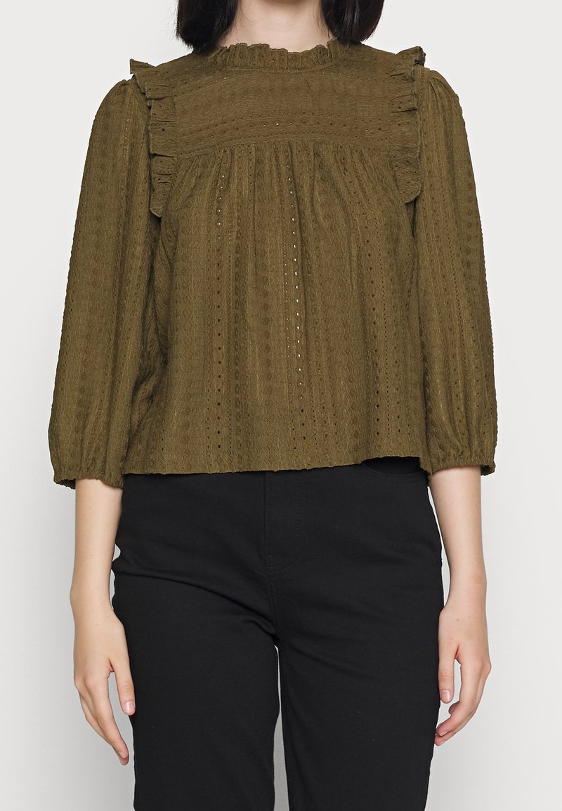 Olivgrün Bluse mit Rüschenausschnitt, strukturiertem Stoff, langen Ärmeln und gerafften Details. Kombiniert mit schwarzen Hosen.
