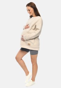 Helles beiges Sweatshirt mit oversized Passform, tiefen Schultern und Logo-Patch. Kombiniert mit grauen Shorts und weißen Sneakers. Das Model hält den Bauch.