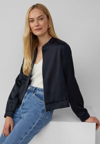 Veste bomber en satin marine avec un col rond, des poignets élastiques et un détail de logo sur le côté, associée à un haut blanc et un jean en denim bleu clair.