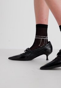 Scarpe in pelle nera con punta affusolata e tacco kitten basso, abbinate a calzini neri a coste con dettagli in catena d'argento.