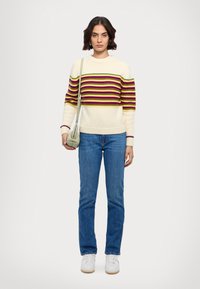 Pull crème à rayures horizontales multicolores, associé à un jean bleu. Le mannequin tient un sac vert clair et porte des baskets blanches.