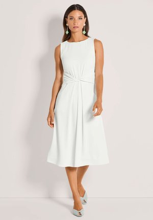 Vrouw draagt een mouwloze witte midi-jurk met gedraaide detail bij de taille, gecombineerd met lichtblauwe open sandalen en geometrische hangende oorbellen.