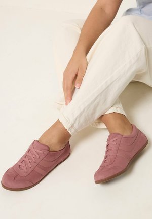 Person seated with crossed legs wearing light pink lace-up sneakers and off-white pants, hand resting on knee.