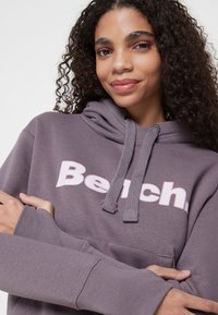 Lila Baumwollkapuzenpullover mit großem pinkem "Bench."-Logo auf der Vorderseite, ausgestattet mit einer Kordelzugkapuze und langen Ärmeln in lockerer Passform.