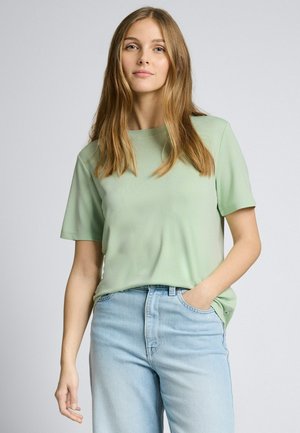 Jeune femme aux cheveux blonds longs portant une chemise à manches courtes vert clair et un jean bleu clair, debout avec une main dans la poche.