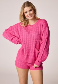 Fényes rózsaszín, kötött, oversized pulóver bordázott mandzsettával és zsebből, azonos anyagú kötött sorttal párosítva. Sima textúra, laza fazon.