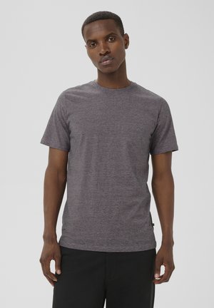 Muž v krátkym rukávom sivom tričku a čiernych nohaviciach stojaci pred jednoduchým svetlosivým pozadím.