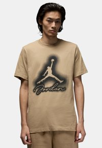 T-shirt in cotone beige caratterizzata da un grande logo silhouette in bianco e nero di Jordan con il testo "Jordan" sottostante, maniche corte e scollatura tonda.