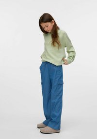 Suéter de punto verde, pantalones cargo azules holgados con bolsillos, pantuflas beiges. Pose de pie, mirando hacia abajo, con cabello largo. Fondo simple.