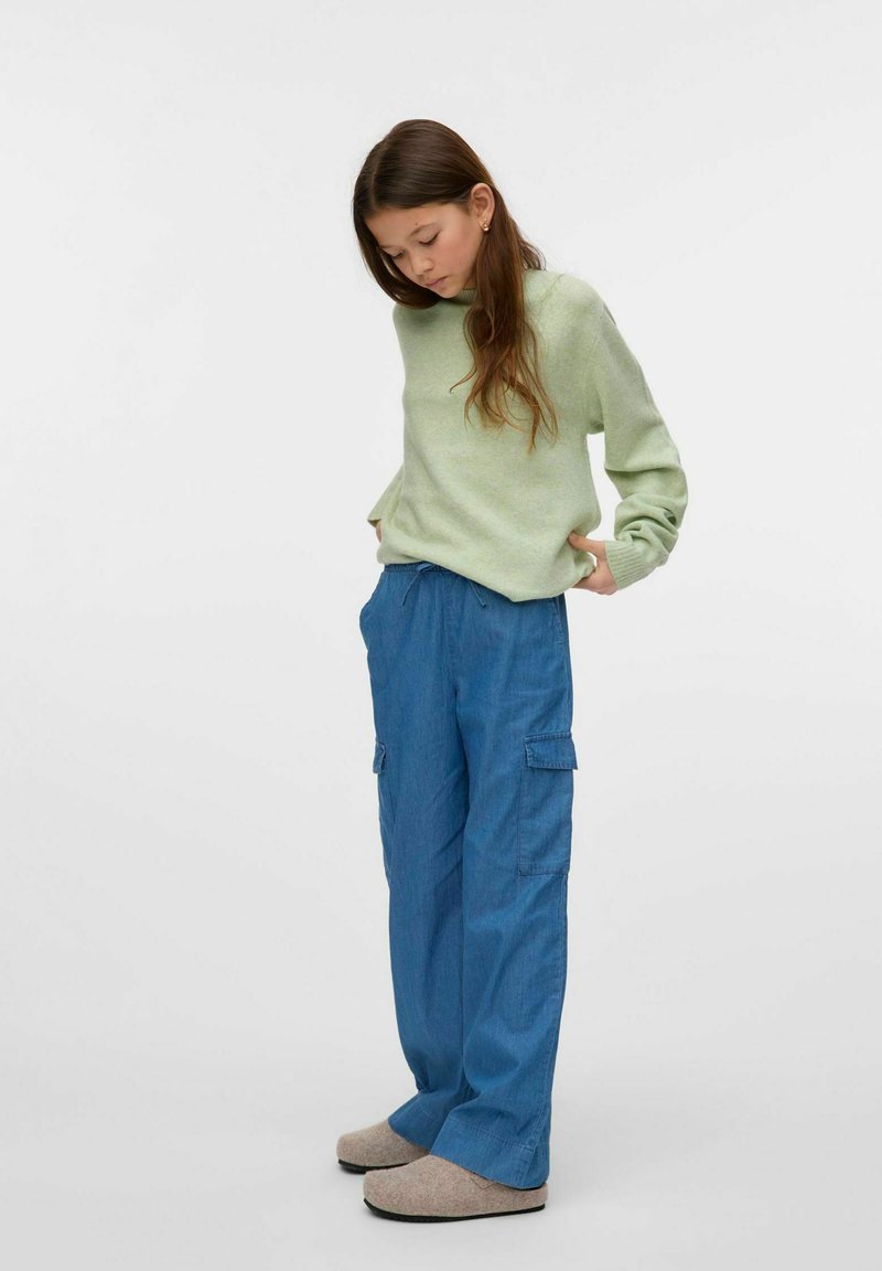 Suéter de punto verde, pantalones cargo azules holgados con bolsillos, pantuflas beiges. Pose de pie, mirando hacia abajo, con cabello largo. Fondo simple.