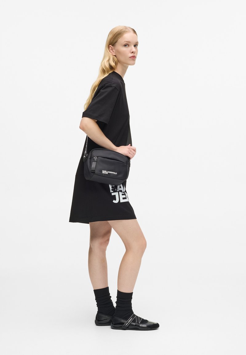 Tricou negru supradimensionat cu text grafic alb, asortat cu o geantă crossbody neagră. Șosete și pantofi negri completează ținuta.