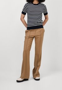 Námořnicky modré a bílé proužkované pletené tričko s krátkým rukávem spojené s hnědými kalhotami s vysokým pasem a krémovými mokasínami, stojící před neutrálním pozadím.