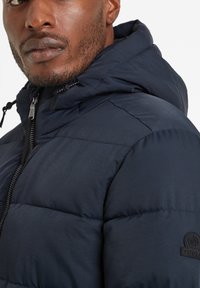 Close-up of a man wearing a zipped dark blue puffer jacket with a hood and a TOG24 logo on the sleeve.