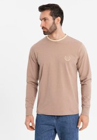 Uomo che indossa una camicia beige a maniche lunghe con un piccolo logo "EX" e colletto a strisce leggere, abbinata a jeans blu, che guarda verso sinistra.