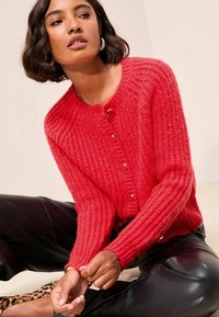 Cardigan rosso lavorato a maglia con texture a coste, scollo rotondo e chiusure a bottoni. Indossato con pantaloni in pelle nera.