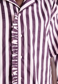 Camisa às riscas em roxo e rosa, com um placket franzido, mangas curtas e botões rosa. O material parece suave e leve.