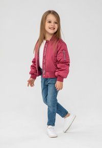 Chaqueta bomber de satén rosa con puños acanalados, combinada con jeans azul claro y zapatillas blancas. El niño tiene el cabello largo y sonríe.