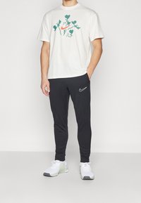 Camiseta blanca de algodón con un logo de Nike en píxeles verdes y naranjas, combinada con pantalones de chándal negros con el swoosh blanco de Nike. Zapatillas deportivas blancas.