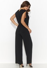 Czarny kombinezon o szerokich nogawkach i drapowanej tylnej części. Wykonany z gładkiego materiału, z głębokim dekoltem w kształcie litery V i delikatnymi fałdami na ramionach.