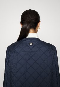 Giacca trapuntata blu navy con scollo rotondo, motivo a diamante e una piccola etichetta nera con un emblema di farfalla bianca sul retro.