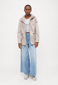 Veste à capuche gris clair avec cordons de serrage, poches avant et doublure beige. Associée à un jean bleu à jambes larges et des baskets grises.