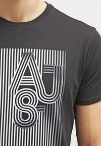 Černé bavlněné tričko s grafickým designem, na kterém jsou tučné bílé pruhované nápisy "AJ" a "8" v různých tloušťkách, které ostře kontrastují s látkou.