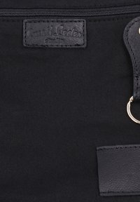 Schwarzer Stoff mit einem aufgenähten Lederlabel mit der Aufschrift "Lusti Leder Studio" und einem schwarzen Lederband mit einem Metallring daran.