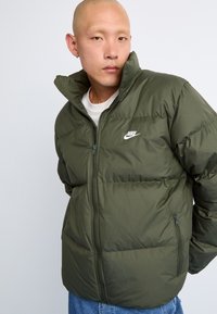Green puffer jacket with a zip front, high collar, and side pockets. Features a white Nike logo on the chest and quilted texture.