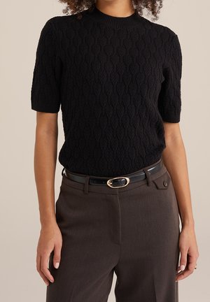 Persoon die een zwarte kabelgebreide trui met korte mouwen draagt, ingestopt in donkergroene, hooggetailleerde broek met een zwarte riem.