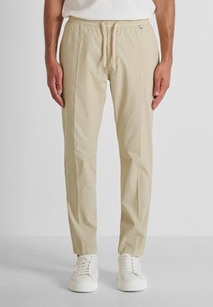 Pantalones casual beige con cintura elástica y cordón, usados con zapatillas blancas y una camisa blanca, mostrados de la cintura a los pies vista frontal.
