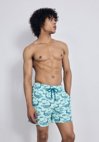 Jeune homme aux cheveux bouclés portant un short de bain bleu clair à motifs de baleines bleu foncé, se tenant les mains dans les poches sur un fond uni.