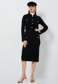 Svart outfit med en figurnära kjol och en skjorta med knäppning och guldaccenter, två bröstfickor och krage. Modellen bär en svart keps och guldskor.