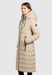 Manteau beige matelassé avec capuche, fermeture zippée à l'avant et poches latérales. Présente une texture matelassée et des accents en métal noir. Design long.