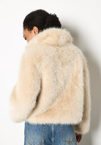 Faux bontjas in lichtbeige met een zachte, textuurafwerking, voorzien van een staande kraag en een cropped zoom, gecombineerd met blauwe denim jeans.