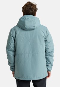 Chaqueta aislante azul claro con capucha, que presenta puños elásticos, una textura suave y un diseño con la parte trasera más larga para mayor cobertura.
