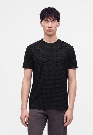 Giovane uomo in piedi che indossa una t-shirt nera con un piccolo motivo "AX" ripetuto e pantaloni grigio scuro su uno sfondo bianco semplice.