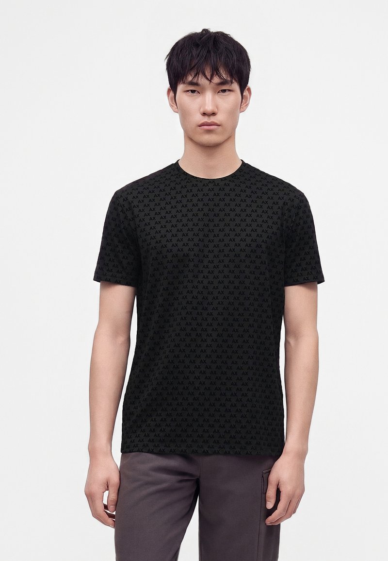 Jeune homme debout portant un t-shirt noir avec un petit motif répétitif "AX" et un pantalon gris foncé sur un fond blanc uni.