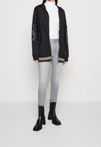 Černá mikina s kapucí s krajkovými panely, pruhovaným lemem a zlatými detaily. Kombinována se světle šedými džínami a černými kotníkovými botami na podpatku.