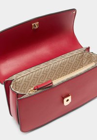 Červená kožená crossbody taška se zlatým čtvercovým zapínáním. Interiér obsahuje světle vzorovanou podšívku a zipovou kapsu.