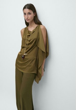Femme portant un haut sans manches asymétrique vert olive et un pantalon large assorti, accessoirisée d'un long collier pendentif, debout contre un mur blanc.