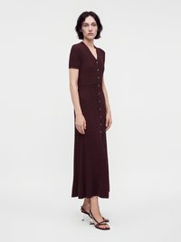 Femme debout portant une robe midi boutonnée bordeaux foncé à manches courtes et des talons noirs à lanières avec talons blocs transparents.