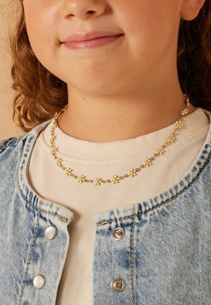 Collier floral en or avec de petites marguerites blanches et jaunes, porté sur un sweat-shirt crème sous une veste en denim claire.